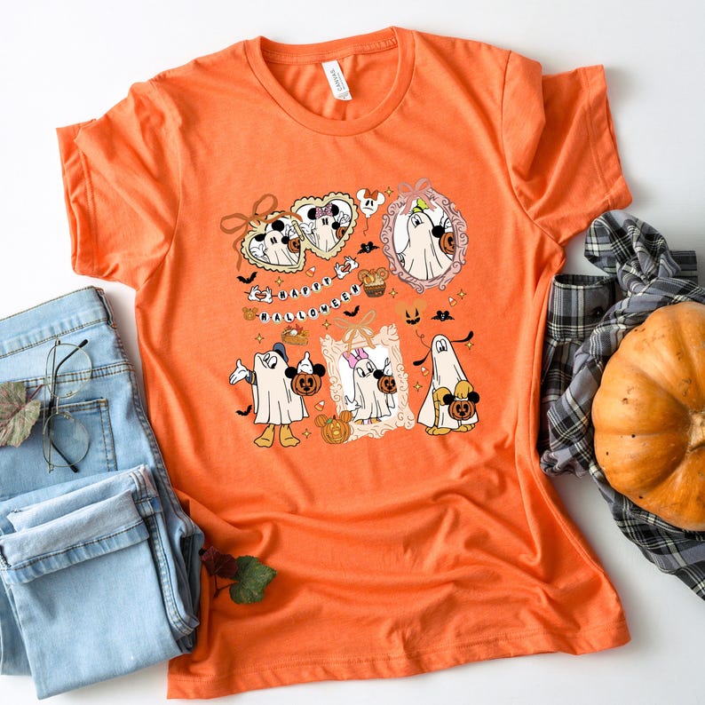 May include: Orange t-shirt with a Halloween-themed graphic. The graphic features cartoon ghosts, pumpkins, and the text "Happy Halloween." The design includes Mickey and Minnie Mouse characters in ghost costumes. The shirt is paired with blue jeans and a pumpkin.