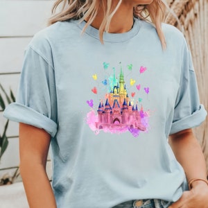 May include: A light blue t-shirt with a watercolor illustration of a pink castle with rainbow Mickey Mouse balloons.