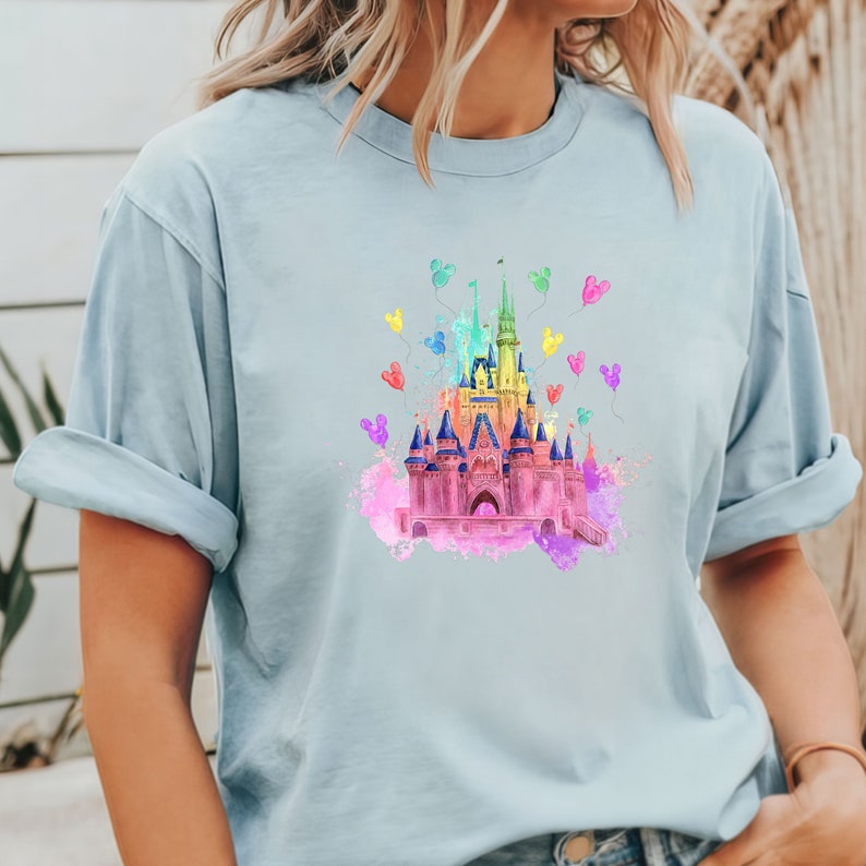 May include: A light blue t-shirt with a watercolor illustration of a pink castle with rainbow Mickey Mouse balloons.