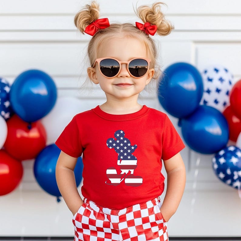 May include: A child in a red t-shirt featuring a patriotic Mickey Mouse design, paired with red and white checkered shorts and round sunglasses. Red bows are in the child's pigtails. Blue, white, and red balloons are in the background.