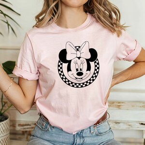 May include: A pink t-shirt featuring a black and white checkered circle surrounding a black and white image of Minnie Mouse with a large bow.