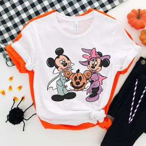 May include: White t-shirt with a graphic of Mickey Mouse and Minnie Mouse dressed in Halloween costumes. Mickey is wearing a vampire costume and Minnie is wearing a witch costume. They are holding a jack-o-lantern.