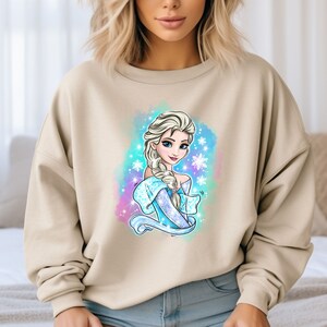Op de afbeelding: Een beige sweatshirt met een kleurrijke illustratie van een cartoonprinses met lang blond haar en een blauwe jurk. De prinses wordt omringd door sneeuwvlokken en een lichtblauwe achtergrond.