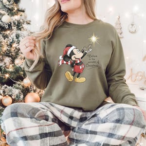 May include: Olive green sweatshirt featuring a graphic of Mickey Mouse in a Santa hat and scarf, pointing at a star. The text "Believe in the wonder of Christmas" is also on the sweatshirt. The person is wearing plaid pajama pants.