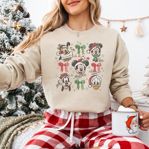 May include: Beige crewneck sweatshirt featuring a festive design with Mickey Mouse and friends in Santa hats and bows. The graphic includes Pluto, Mickey, Minnie, Goofy, and Donald Duck. A Christmas-themed apparel item.