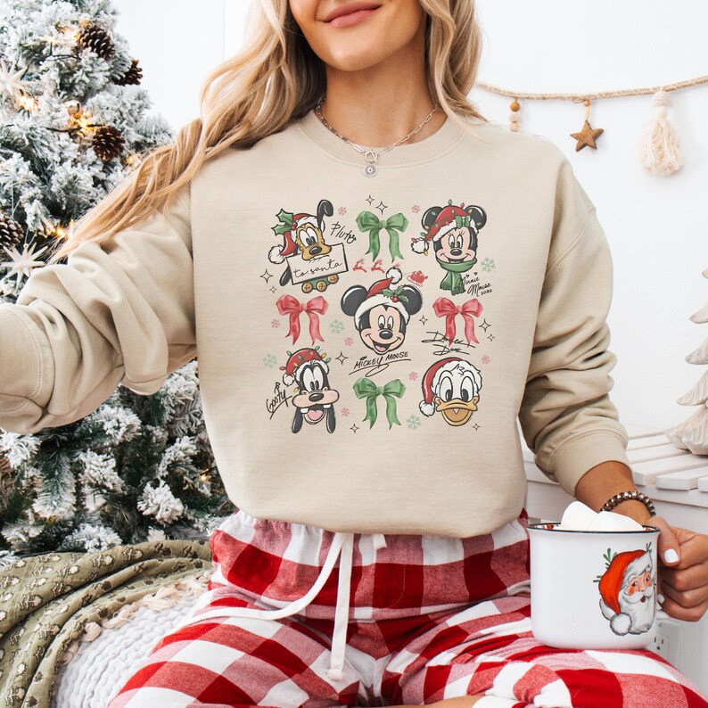 May include: Beige crewneck sweatshirt featuring a festive design with Mickey Mouse and friends in Santa hats and bows. The graphic includes Pluto, Mickey, Minnie, Goofy, and Donald Duck. A Christmas-themed apparel item.