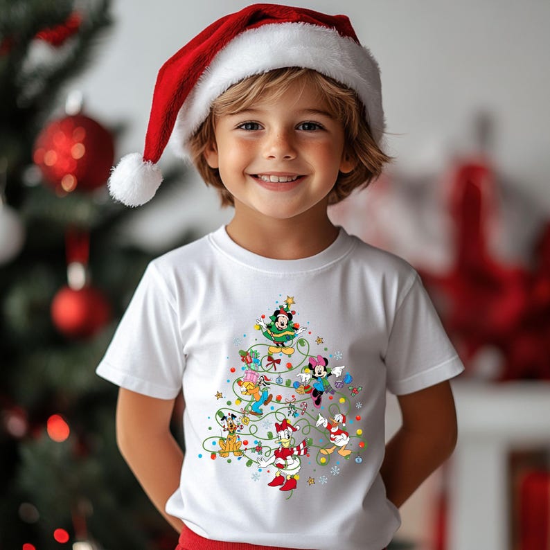 K&ouml;nnte beinhalten: Ein Kind tr&auml;gt ein wei&szlig;es T-Shirt mit einem Weihnachtsbaum-Design mit Disney-Figuren. Das Kind tr&auml;gt auch einen roten Weihnachtsmannhut mit einem wei&szlig;en Bommel. Im Hintergrund ist ein geschm&uuml;ckter Weihnachtsbaum zu sehen.