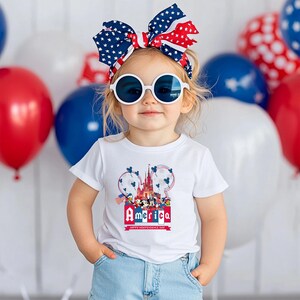 Może przedstawiać: Dziecko w białej koszulce z kolorową grafiką "America" przedstawiającą postacie Disneya i zamek. Dziecko ma r&oacute;wnież na sobie patriotyczną kokardę, okulary przeciwsłoneczne i dżinsowe szorty. Czerwone, białe i niebieskie balony w tle.