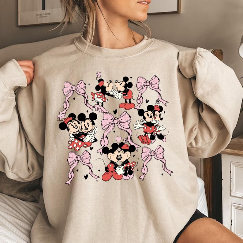May include: A beige sweatshirt with a graphic print of Mickey Mouse and Minnie Mouse in various poses. The print features pink bows and hearts.