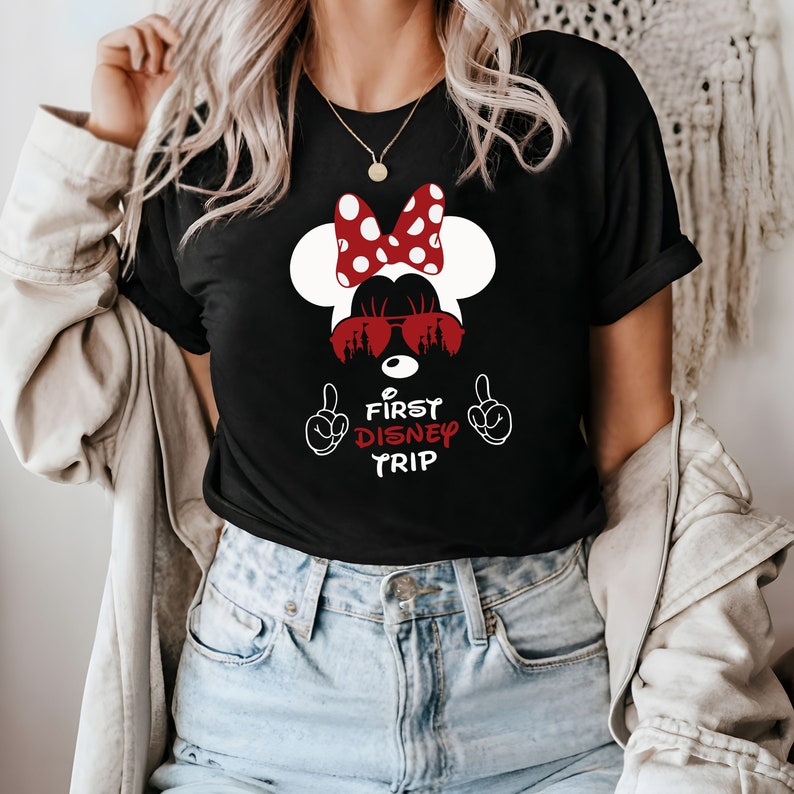 Op de afbeelding: Zwart T-shirt met een witte en rode Minnie Mouse-afbeelding. De afbeelding toont Minnie Mouse met een zonnebril waarin het Disney-kasteel wordt weerspiegeld. De tekst "First Disney Trip" staat onder de afbeelding.