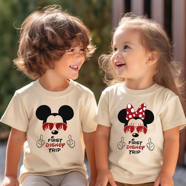 Op de afbeelding: Twee kinderen dragen cr&egrave;mekleurige T-shirts met een Mickey Mouse en Minnie Mouse afbeelding. De shirts zeggen "First Disney Trip" met een rode kasteelafbeelding in de zonnebril.