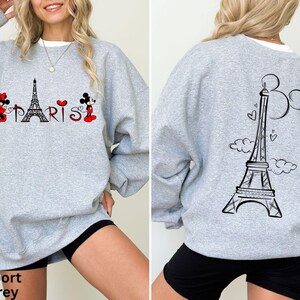 K&ouml;nnte beinhalten: Hellgraues Sweatshirt mit einem Front-Design, das den Eiffelturm, das Wort "PARIS" und Zeichentrickfiguren zeigt. Die R&uuml;ckseite des Sweatshirts hat ein schwarzes Eiffelturm-Design mit Herzen und Wolken. Der Text "Sport Grey" steht unten.
