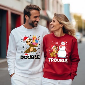 Puede incluir: Una sudadera blanca con un dibujo animado de una ardilla llevando regalos y el texto "Double Trouble". Una sudadera roja con un dibujo animado de una ardilla construyendo un mu&ntilde;eco de nieve y el texto "Trouble".