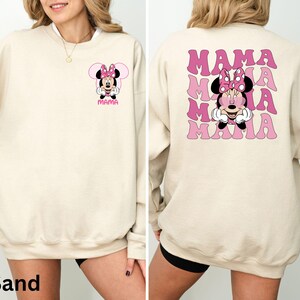 Puede incluir: Sudadera color arena con un gr&aacute;fico de Minnie Mouse, lazo rosa y la palabra "MAMA". La parte trasera presenta un gr&aacute;fico de Minnie Mouse m&aacute;s grande y la palabra "MAMA" en rosa, con fuente ondulada. Un estilo casual y c&oacute;modo.