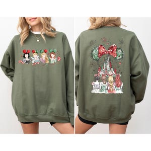 May include: Olive green sweatshirt featuring a watercolor design of Disney princesses with holiday-themed mouse ears and candy canes. The back of the sweatshirt showcases a castle and more princesses, with a large red bow and fireworks.