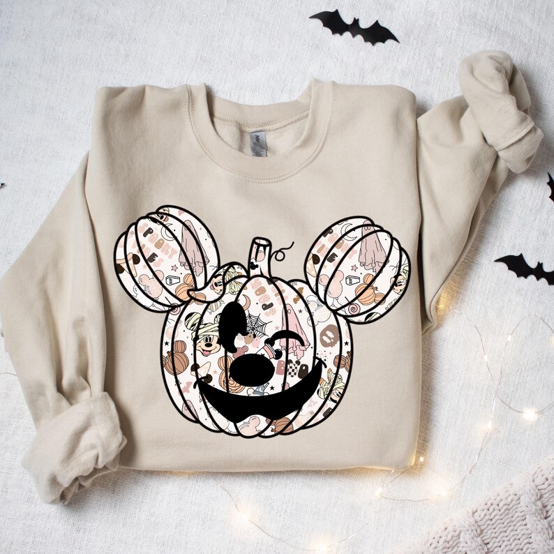 Op de afbeelding: Een beige sweatshirt met een zwart-wit pompoenontwerp. De pompoen is gemaakt van kleinere afbeeldingen van Halloween-thema's, waaronder geesten, vleermuizen en ma&iuml;s snoepjes. De pompoen heeft Mickey Mouse-oren.