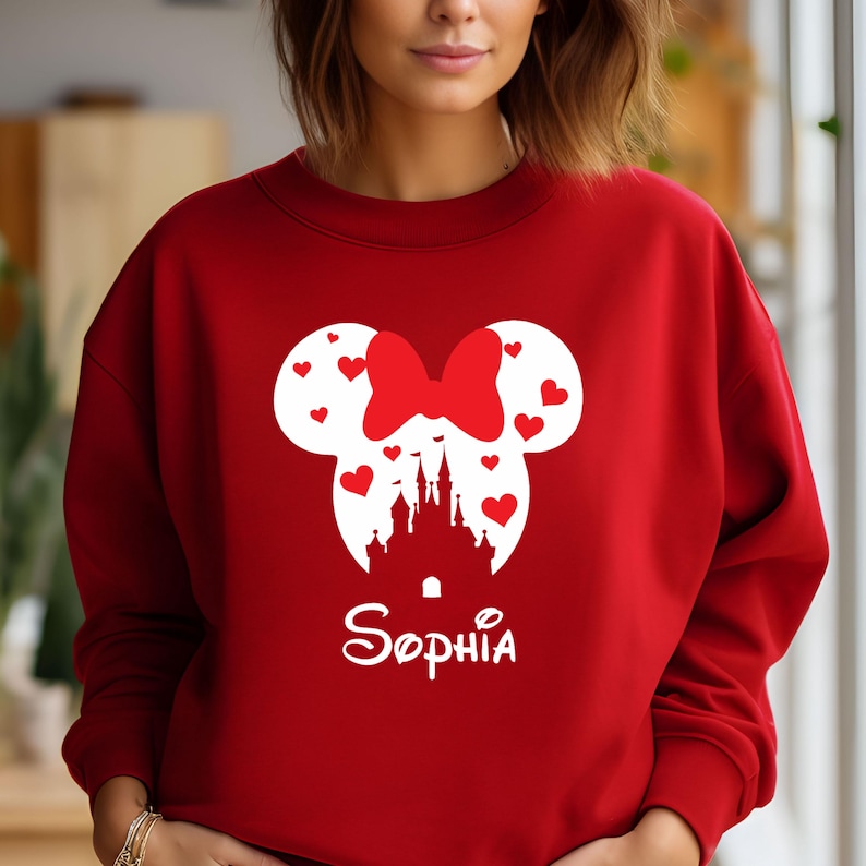 May include: Red sweatshirt featuring a white silhouette of Mickey Mouse ears with a bow and a castle inside. The name "Sophia" is written below the silhouette.