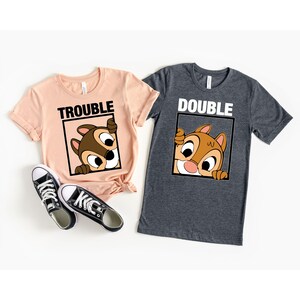 May include: Two t-shirts with a cartoon chipmunk peeking out of a box. The pink shirt says "Trouble" and the gray shirt says "Double".