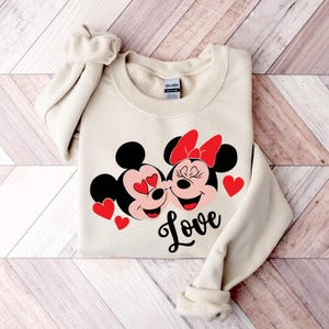 May include: A beige sweatshirt with a graphic of Mickey Mouse and Minnie Mouse kissing. The graphic is in red, black, and white. The text "Love" is printed below the graphic.