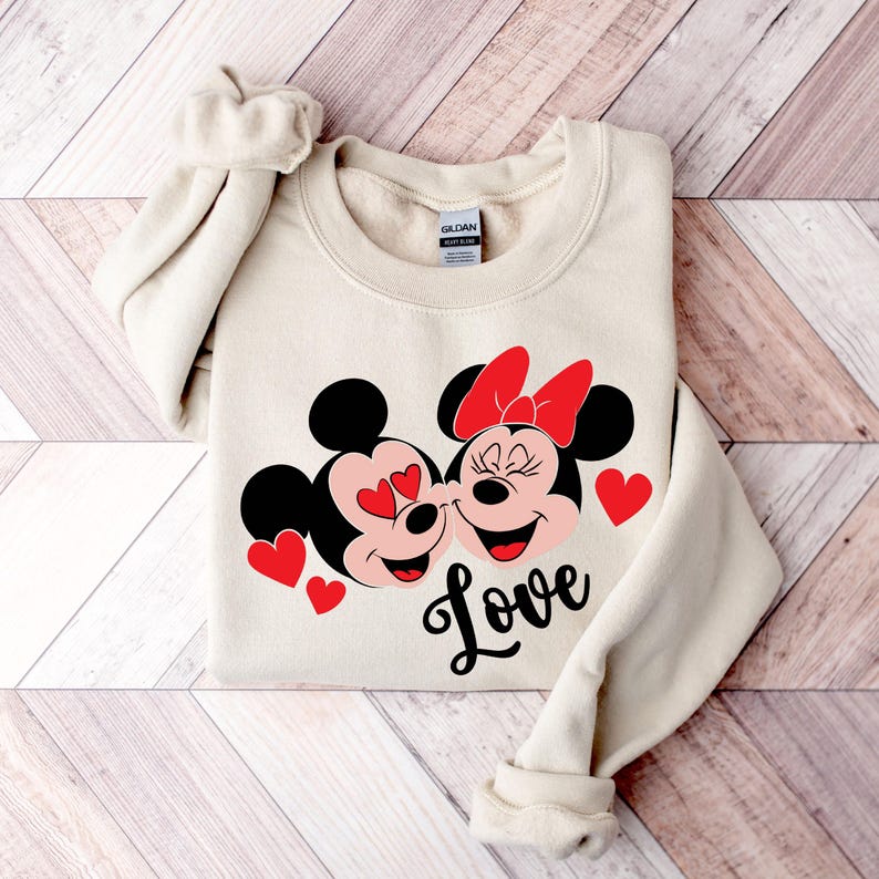 May include: A beige sweatshirt with a graphic of Mickey Mouse and Minnie Mouse kissing. The graphic is in red, black, and white. The text "Love" is printed below the graphic.