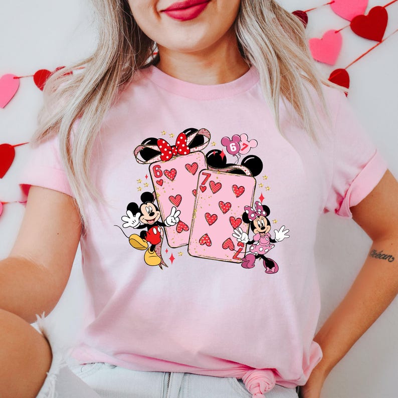 May include: Pink t-shirt featuring Mickey and Minnie Mouse with playing card designs. The cards have heart patterns and the numbers 6 and 7. Mickey Mouse is wearing red shorts and Minnie Mouse is wearing a pink dress.