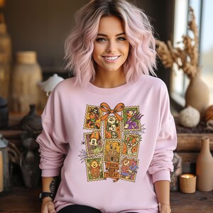 May include: A light pink sweatshirt featuring a Halloween-themed graphic. The graphic includes postage stamp-style images of cartoon characters and a haunted house, with the words "Trick or Treat". The sweatshirt has long sleeves and a crew neck.