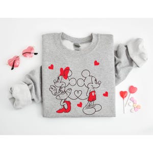 Op de afbeelding: Een grijs sweatshirt met een afbeelding van Mickey Mouse en Minnie Mouse die hand in hand staan. De afbeelding is in zwart en rood met hartjes eromheen.