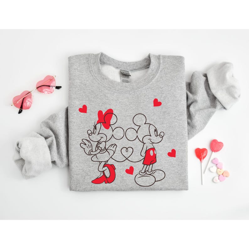 Op de afbeelding: Een grijs sweatshirt met een afbeelding van Mickey Mouse en Minnie Mouse die hand in hand staan. De afbeelding is in zwart en rood met hartjes eromheen.
