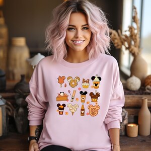 May include: A light pink sweatshirt featuring a collection of cartoon food and drink icons. The icons include a Mickey Mouse-shaped ice cream bar, donut, cupcake, coffee, and more. The design has a fall theme with autumn leaves.