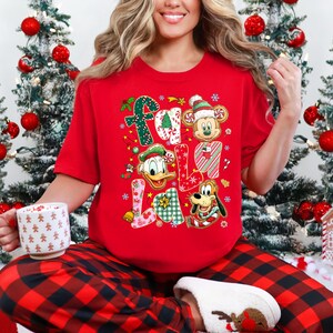 K&ouml;nnte beinhalten: Rotes Weihnachtsshirt mit den Worten "Fa La La" in einem farbenfrohen, festlichen Design. Das Shirt zeigt Bilder von Mickey Mouse, Donald Duck und Pluto. Die Person tr&auml;gt eine rot-schwarz karierte Pyjamahose und wei&szlig;e Hausschuhe.