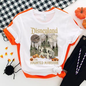 May include: White t-shirt with a vintage-style graphic of Disneyland's Haunted Mansion. The graphic features Mickey Mouse and a ghost holding a book that says "Trick or Treat". The text "Disneyland" and "The Haunted Mansion" are in gold lettering.