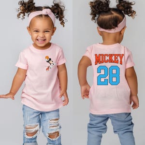 May include: Pink short-sleeved t-shirt with a cartoon image of Mickey Mouse playing basketball on the front. The back of the shirt has the text "MICKEY 28" in blue and orange.