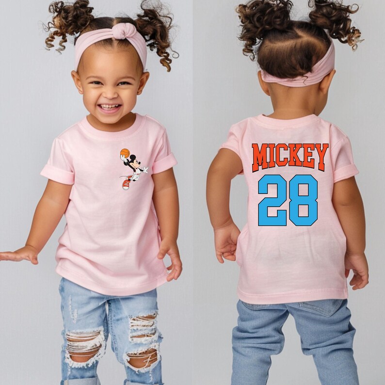 May include: Pink short-sleeved t-shirt with a cartoon image of Mickey Mouse playing basketball on the front. The back of the shirt has the text "MICKEY 28" in blue and orange.