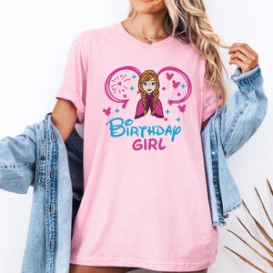May include: Pink t-shirt with a graphic of a cartoon character with a pink cape and blonde hair. The graphic also includes Mickey Mouse ears, fireworks, and the text "Birthday Girl".