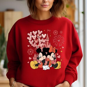 May include: Red sweatshirt with a graphic of Mickey and Minnie Mouse in front of Cinderella's Castle with balloons and fireworks. The text on the balloons says "LOVE" and "XOXO".