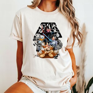 May include: A white t-shirt with a black and white graphic of Mickey Mouse, Donald Duck, and Minnie Mouse dressed as Star Wars characters. The graphic includes the Star Wars logo and a black and white background.