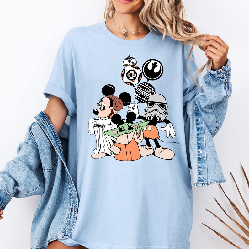 May include: Light blue t-shirt featuring a graphic of Mickey and Minnie Mouse, Baby Yoda, and a Stormtrooper. The design includes Star Wars themed balloons. The shirt is a comfortable, casual style.
