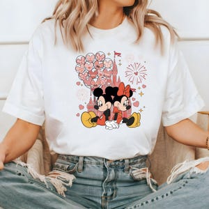 May include: White t-shirt with a graphic of Mickey Mouse and Minnie Mouse holding hands in front of a pink castle with pink balloons and fireworks.