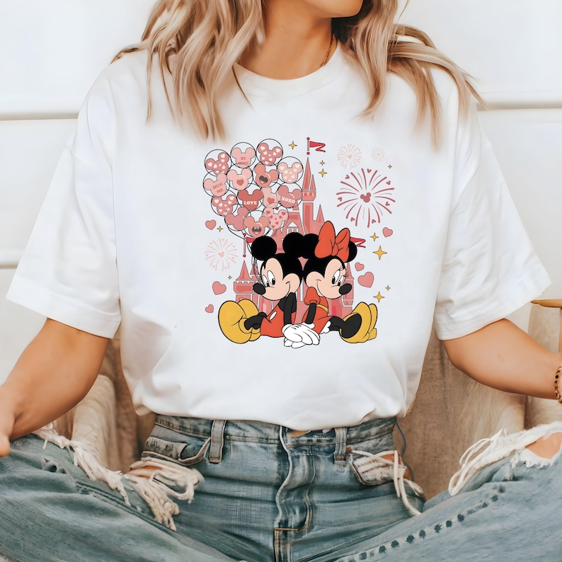 May include: White t-shirt with a graphic of Mickey Mouse and Minnie Mouse holding hands in front of a pink castle with pink balloons and fireworks.