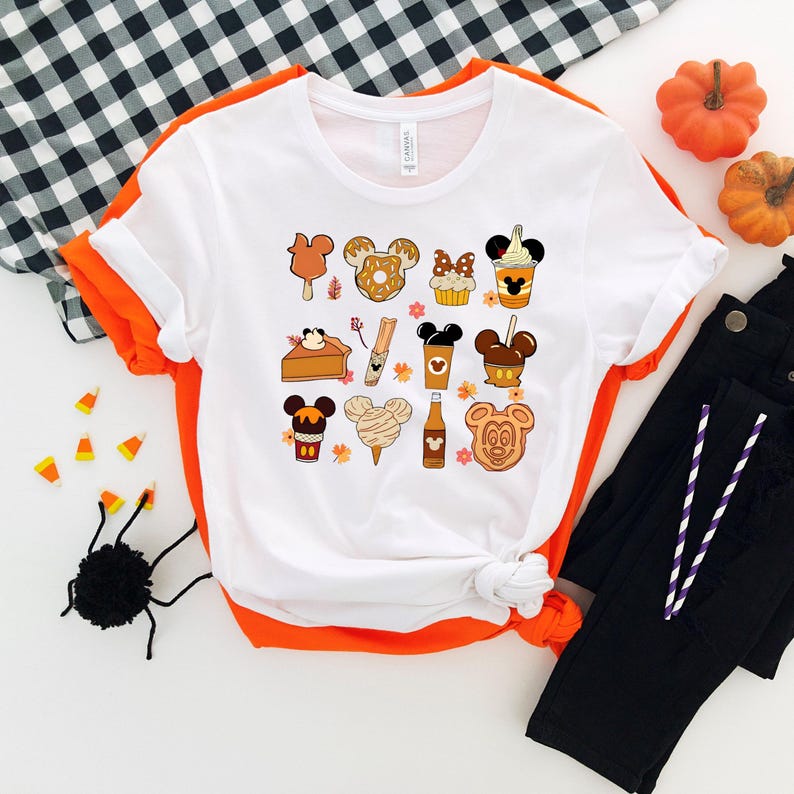 May include: White t-shirt featuring a collection of cartoon illustrations of fall-themed treats and Mickey Mouse-inspired designs. The shirt is surrounded by Halloween decorations, including pumpkins, candy corn, and a black spider.