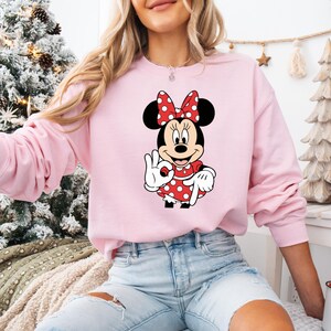 May include: A pale pink sweatshirt with a Minnie Mouse design. The graphic depicts Minnie Mouse in a red polka dot dress and bow, making an 'okay' sign. The sweatshirt is long-sleeved and has a crew neck.