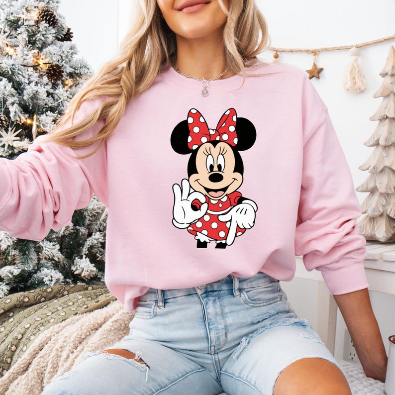 May include: A pale pink sweatshirt with a Minnie Mouse design. The graphic depicts Minnie Mouse in a red polka dot dress and bow, making an 'okay' sign. The sweatshirt is long-sleeved and has a crew neck.