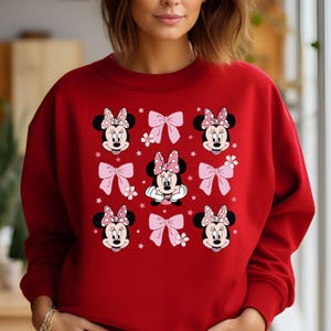 May include: Red sweatshirt with a pattern of Minnie Mouse faces and pink bows. The Minnie Mouse faces are wearing red polka dot bows.