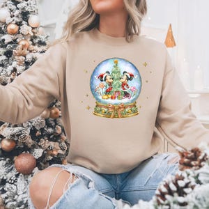 May include: Beige sweatshirt with a snow globe design featuring Mickey and Minnie Mouse in festive Christmas outfits. The snow globe includes a Christmas tree, gifts, and sweets. Gold stars and a bow decorate the base of the globe.