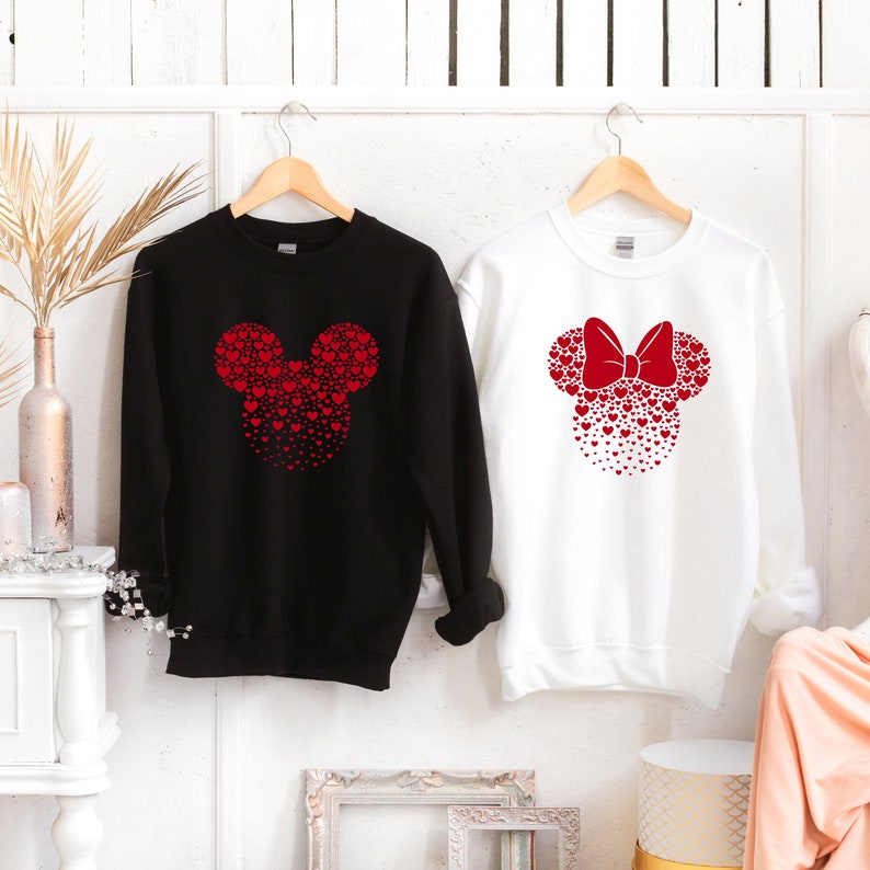 May include: Two black and white sweatshirts with red heart designs. The black sweatshirt has a Mickey Mouse head design made of red hearts. The white sweatshirt has a Minnie Mouse head design made of red hearts with a red bow.