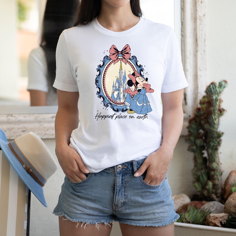 May include: White t-shirt with a graphic of Minnie Mouse standing in front of Cinderella's Castle in a blue and white frame with a pink bow. The text "Happiest place on earth" is printed below the graphic.