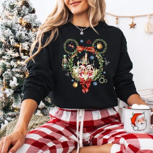May include: Black sweatshirt featuring a Christmas-themed Mickey and Minnie Mouse design. The graphic includes a wreath, castle, and bows. The person is wearing red and white plaid pajama pants and holding a mug with a Santa Claus design.