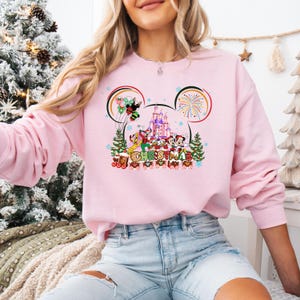 May include: A light pink sweatshirt featuring a festive Christmas design. The graphic includes Mickey Mouse, Minnie Mouse, Donald Duck, and Goofy in front of a castle, with the word "CHRISTMAS" below.