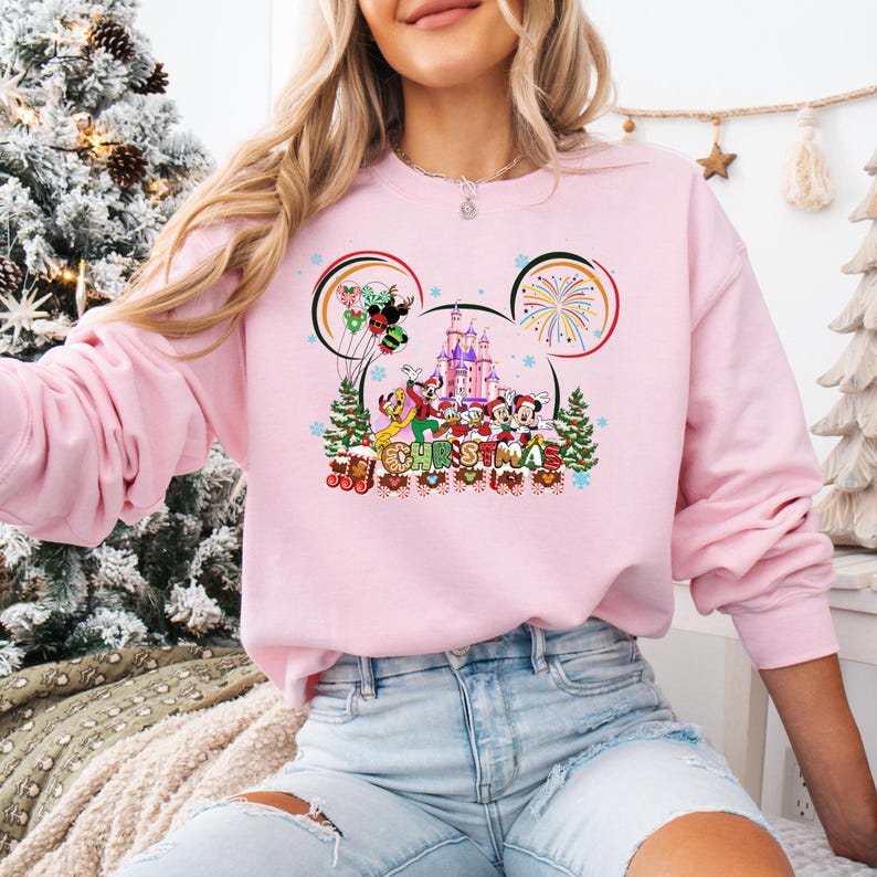 May include: A light pink sweatshirt featuring a festive Christmas design. The graphic includes Mickey Mouse, Minnie Mouse, Donald Duck, and Goofy in front of a castle, with the word "CHRISTMAS" below.