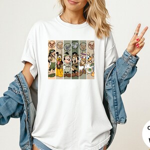 May include: White t-shirt featuring a graphic of Disney characters in safari outfits. The design includes Goofy, Donald Duck, Mickey Mouse, and Minnie Mouse. The shirt has the words "Comfort Colors White" on the bottom right.